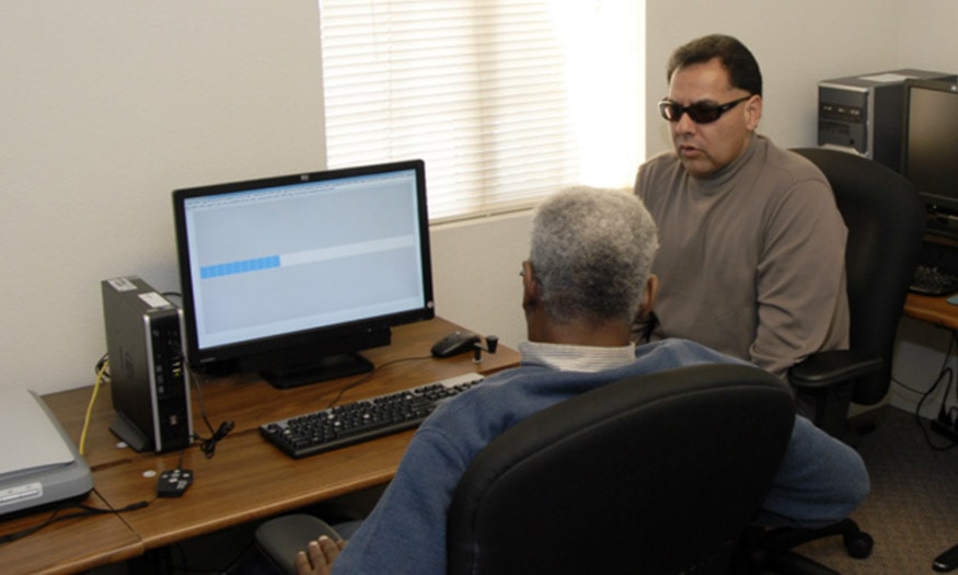 An instructor shows a student how to use a new screen reader technology. Both men are visually impaired.