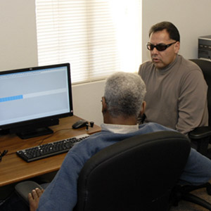 An instructor helps a student learn to use new screen reading technologies. Both men are visually impaired.